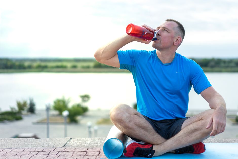 A man in a blue shirt sitting outdoors on an exercise mat, drinking from a water bottle.