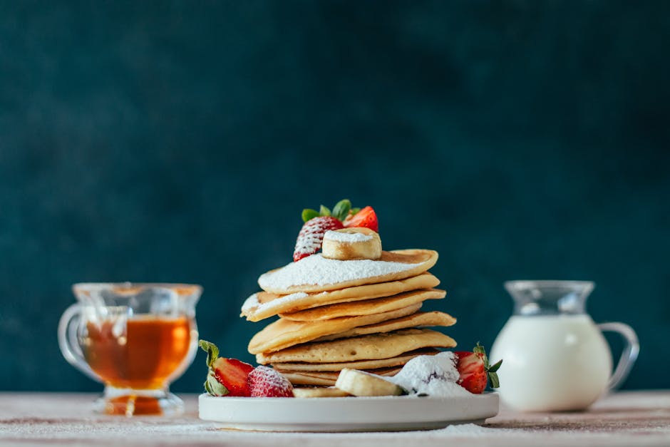 A stack of fluffy pancakes topped with fruits, powdered sugar, honey, and cream.