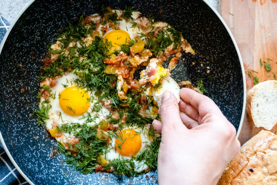 Close-up of homemade shakshuka with fried eggs and herbs in a pan, hand taking a bite.