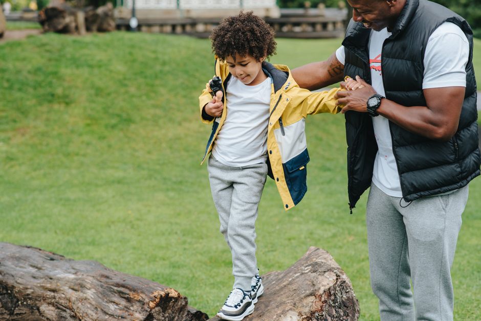 Caring African American man holding hands of careful kid while walking on wooden log in grassy park on blurred background