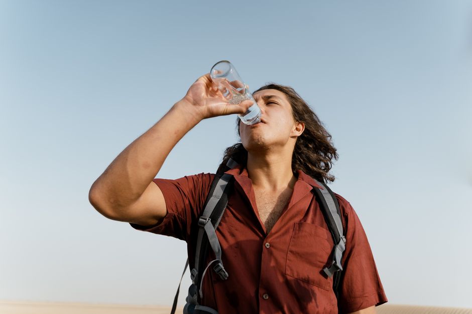 A young man in a red shirt drinks water from a bottle under a clear sky, highlighting hydration and outdoor adventure.