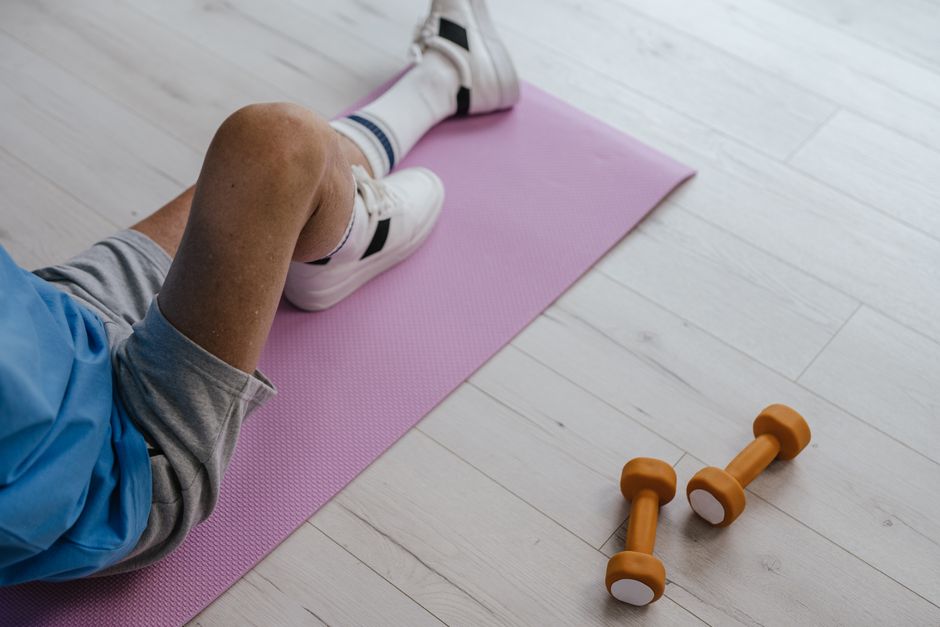 Senior adult sitting on a pink yoga mat indoors with orange dumbbells, promoting a healthy